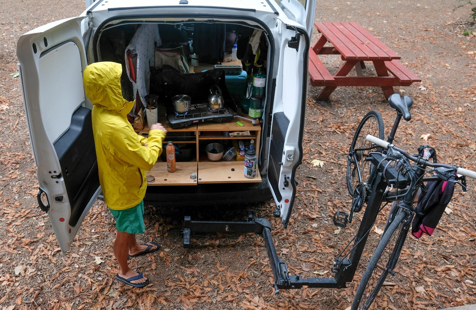 a woman making coffee in a yellow rain coat outside the back of a camper van.
