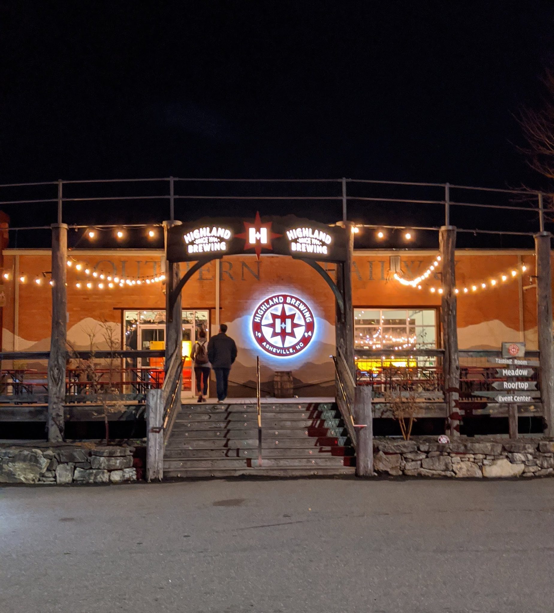 Two people walking into the front doors at Highland Brewing Company at night. There is a sign with the Highland logo and it is lit up.