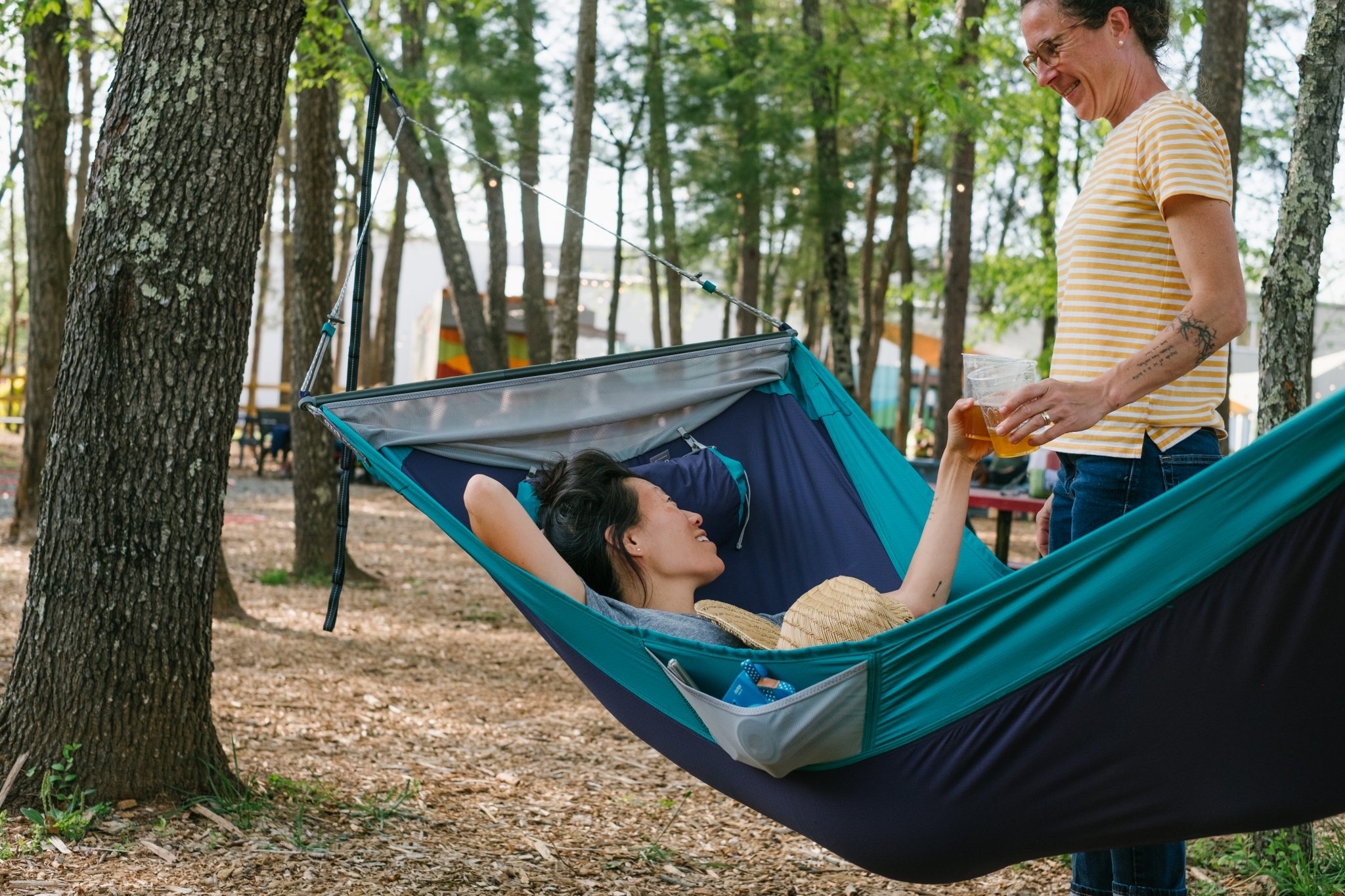 Erin McGrady is laying in a blue hammock toasting Caroline Whatley with a beer. Highland Brewing Company's shipping containers are in the background.