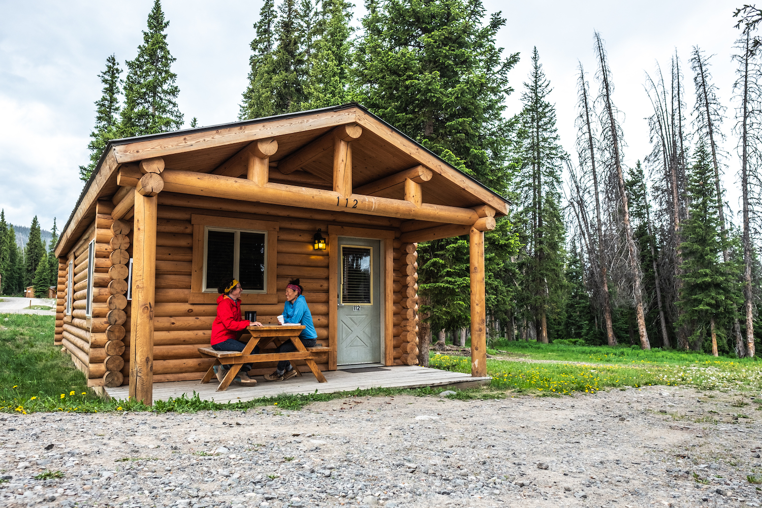 Caroline Whatley and Erin McGrady sit at a picnic table in front of their cabin at Togwotee Mountain Lodge. Caroline is in a red jacket and Erin is in a blue jacket.