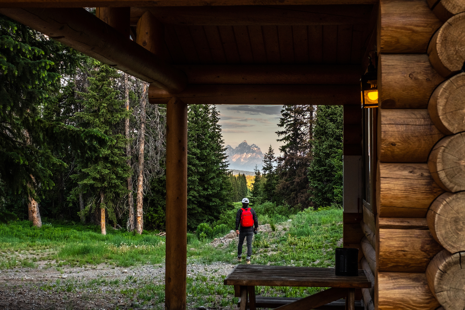 Erin McGrady walks outside the cabin with a camera in hand. The Grand Tetons are in the distance. The cabin and it's wooden slats are in the right hand side of the photo. The porch light on the cabin is on.