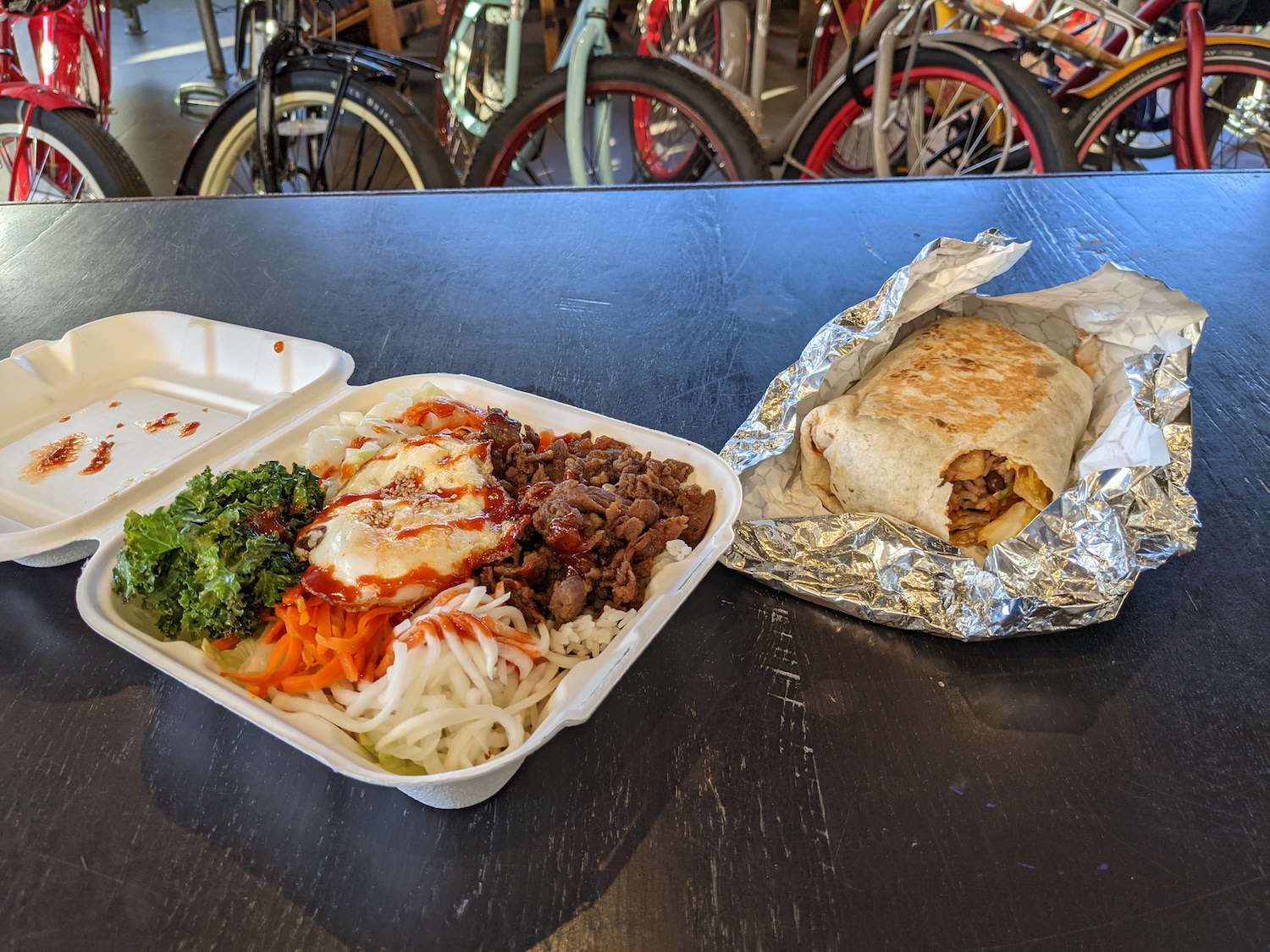 Beef bibimbap is sitting in a container on top of a black table at New Belgium Brewing in Asheville, NC. A burrito is also on the table partially wrapped in foil. Bikes are in the background.
