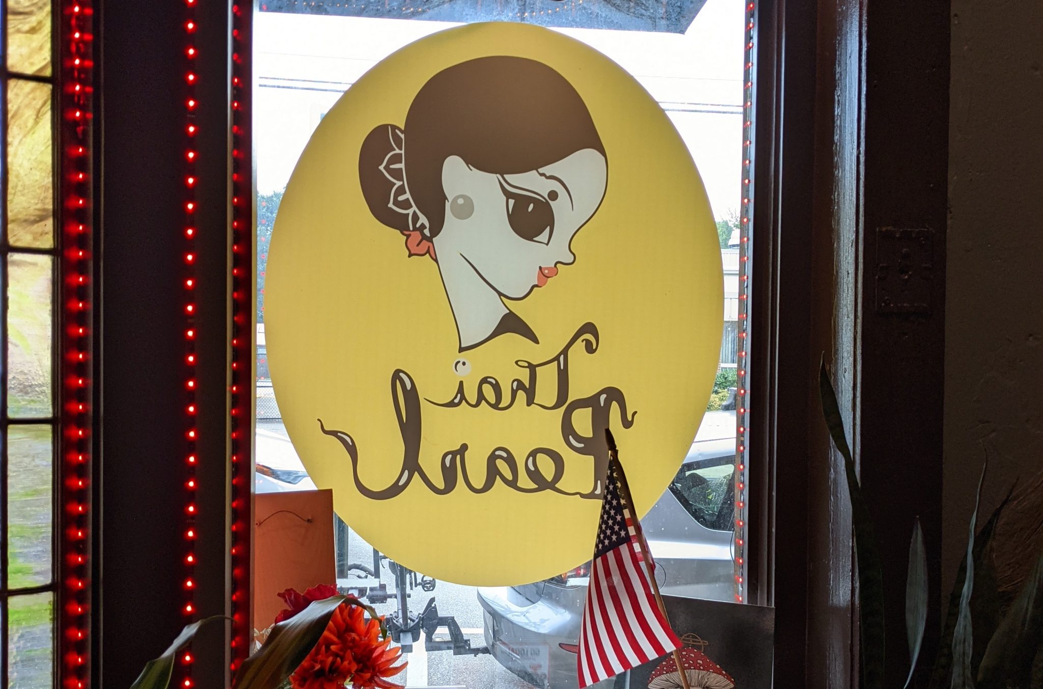 a photo of Thai Pearl's logo painted on their window in West Asheville. The logo, which is yellow with black and white words in cursive, is backwards, as the photograph was taken inside the restaurant. A cartoonish drawing of a woman is above the words Thai Pearl. An American flag is in the foreground and red lights frame the window.