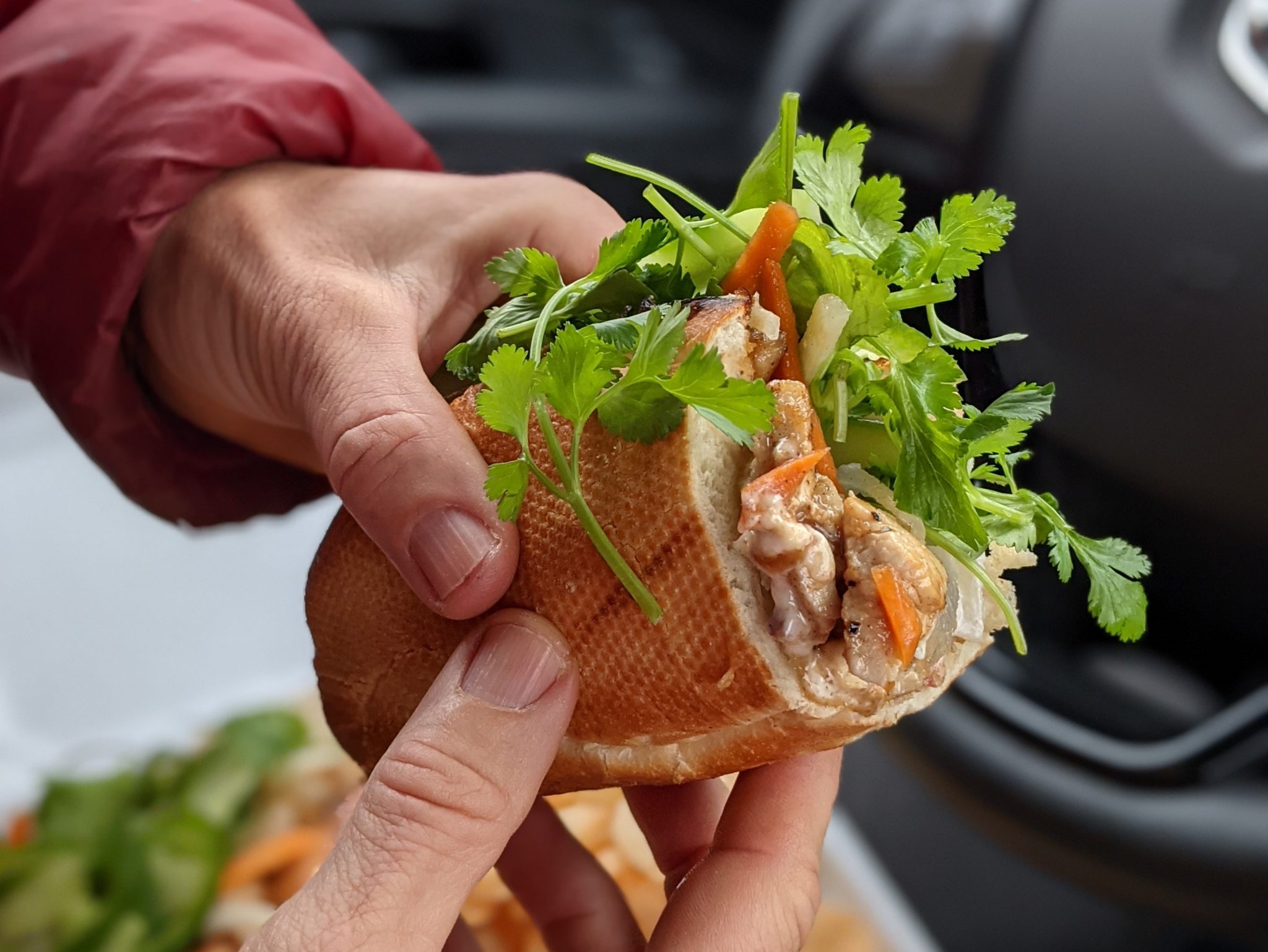 A photo of half of a banh mi sandwich from Wild Ginger is held in Caroline Whatley's hand. A steering wheel and a to-go container is blurred out in the background.