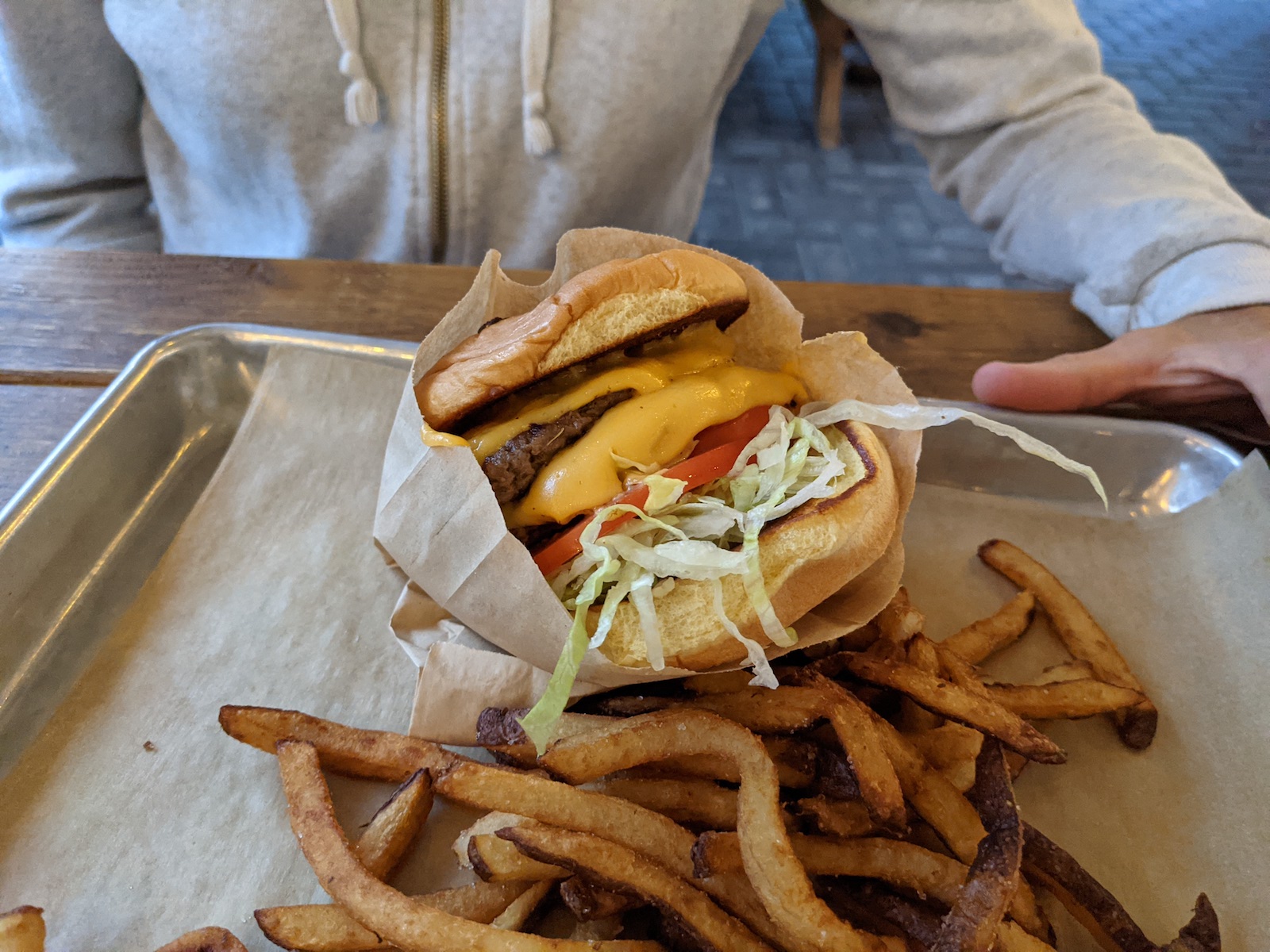 A double patty cheeseburger with a slice of tomato and shredded lettuce is served on a silver tray with a layer of brown paper on the bottom. Fries are on top of the paper and so is the burger which is also enclosed in a tight brown paper bag. A hand rests on the tray which sits on a wooden picnic table.