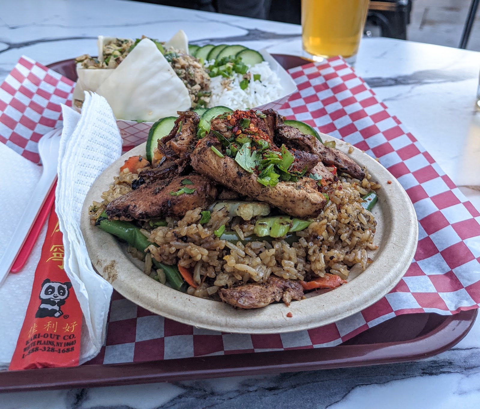 a photo of Thai Fried Rice from Bun Intended is served on a brown paper plate which rests on top of red and white checked paper on top of a brown plastic tray. The tray is on top of a marble table and there are chopsticks and plastic cutlery to the left. A dish of Larb Gai is in the background and so is a glass of beer.