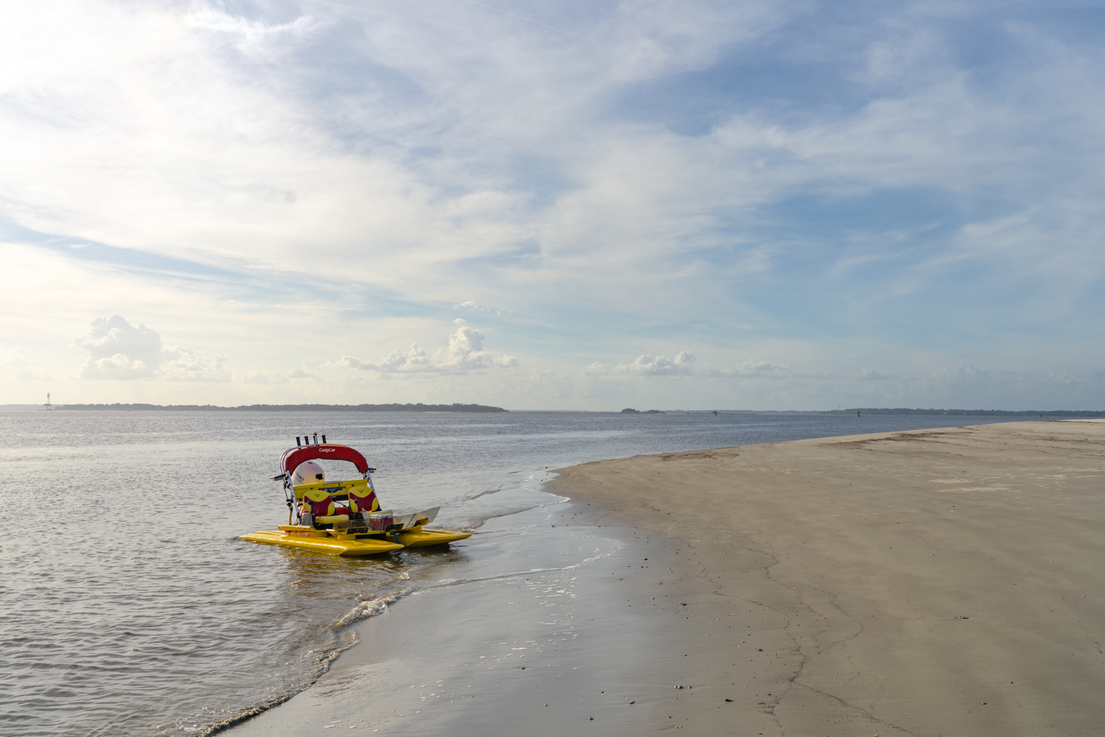A yellow and red CraigCat with a white engine is parked on the shore of a desolate beach. A few clouds are in the background as well as a small strip of land. The sand is untouched and the scene is serene.