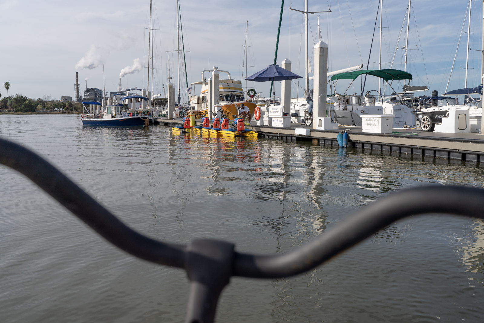 The black handlebars of a River Cycle is blurred out in the foreground. Between the handlebars is a sharp photo of dark water and a dock with nice boats and a blue umbrella, a dock, and four yellow boats with orange life jackets tied to them. A man crouches down next to them. 