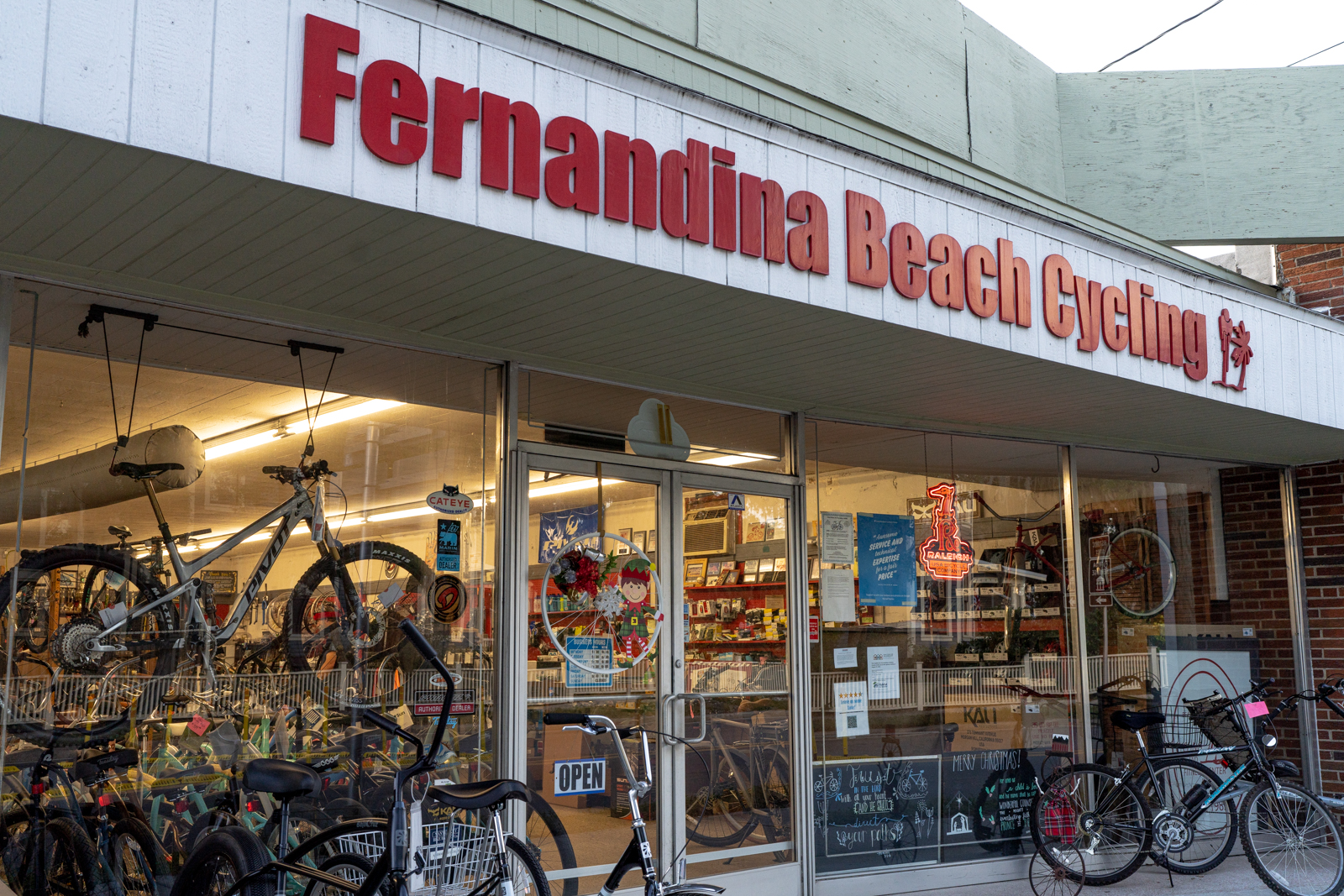 This is a photo of the Fernandina Beach Cycling store. There are bikes in the big glass shop windows and several bikes parked outside. There is an open sign on the door and a Christmas elf as well. Above the glass windows and doors are red letters and the logo of Fernandina Beach Cycling.