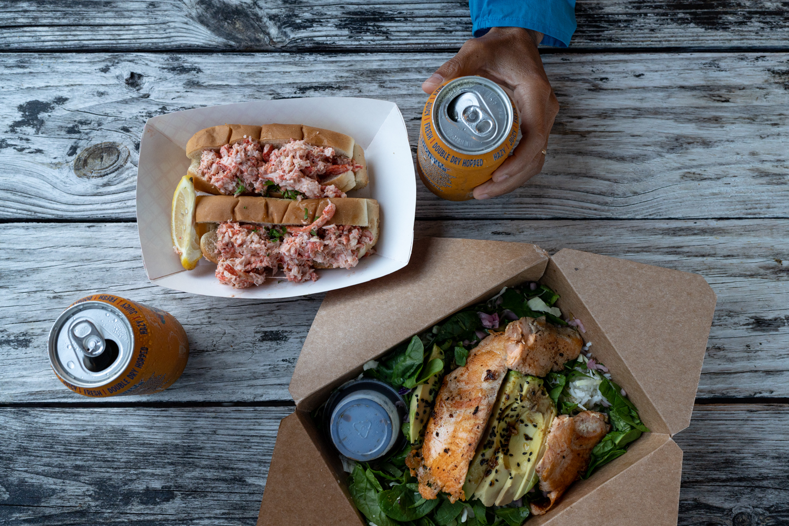 A small hand holds an orange beer can on top of an old wooden picnic table. There is another orange beer can in the bottom left hand corner. There is a white cardboard dish holding two lobster rolls with a lemon wedge and another dish with salad greens, avocado, and grilled salmon in it. A plastic ramakin with dressing is upside down in the same dish.