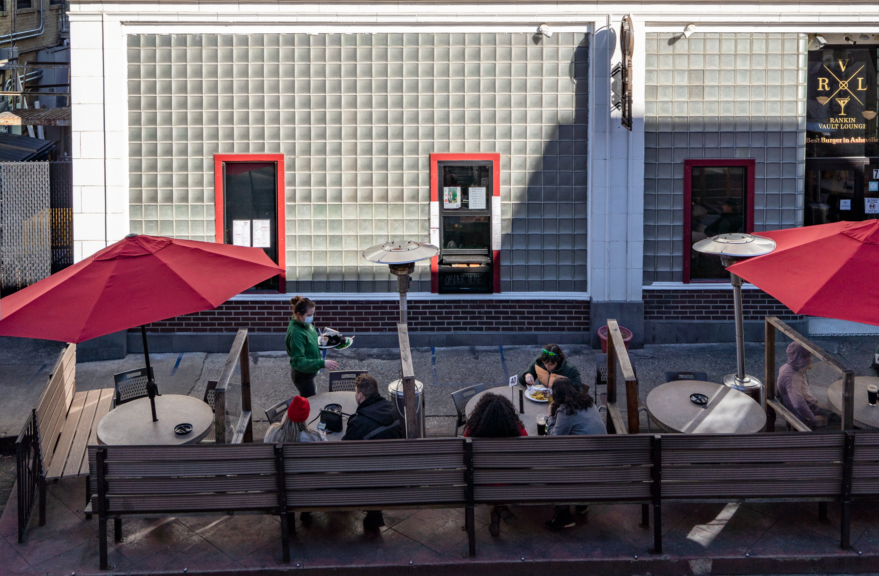 a photo of the outdoor patio of the Rankin Vault Cocktail Lounge. A server in a green shirt holds two dirty plates and approaches a table. Three of the round tables have people in them. There are red umbrellas over most of the tables.