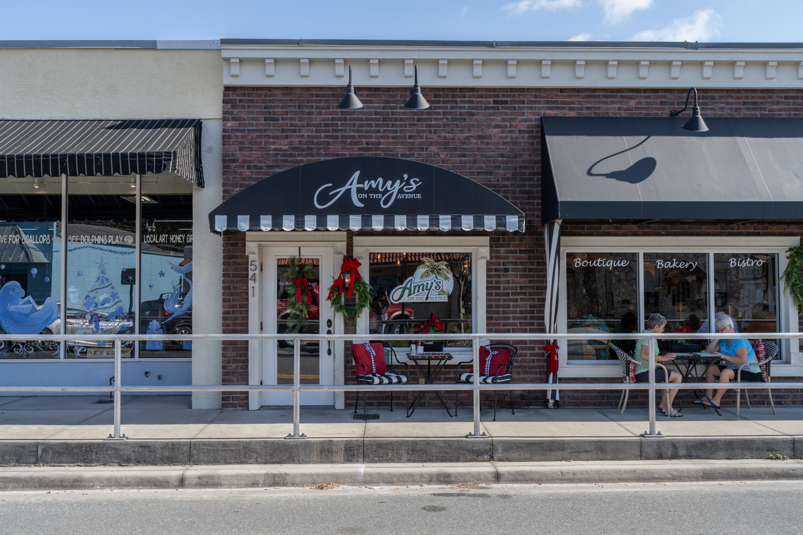 A photo of the exterior and front of Amys on the Avenue in Crystal River. Two people sit at an outdoor table reading a menu. They are facing one another. The entrance is decorated with two green wreaths with a red bow.
