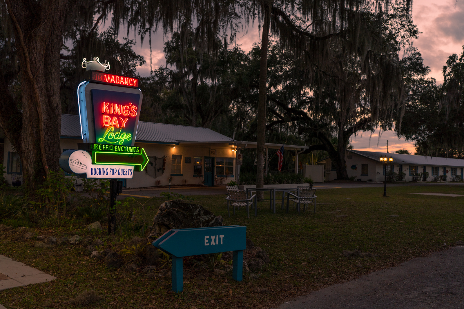The King's Bay Lodge's iconic neon sign is lit up advertising vacancy and 18 efficiency units and docking for guests. Most of the rest of the photo is dark as it is night. There are a few porch lights on in the background, dimly light up the lodge. 
