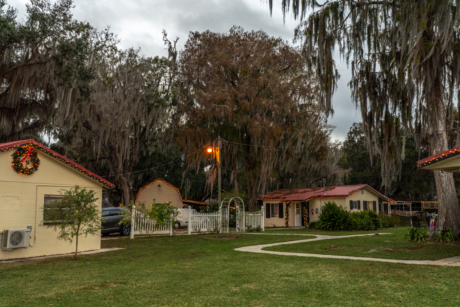 A wide-angle shot of Sunshine Lodge. There are three yellow buildings in the photo, each of which are decorated with yellow Christmas lights. The building on the left has a wreath on the side of it. Spanish moss hangs from trees and there is a light lit up on a short wooden telephone pole in the middle of the photo.