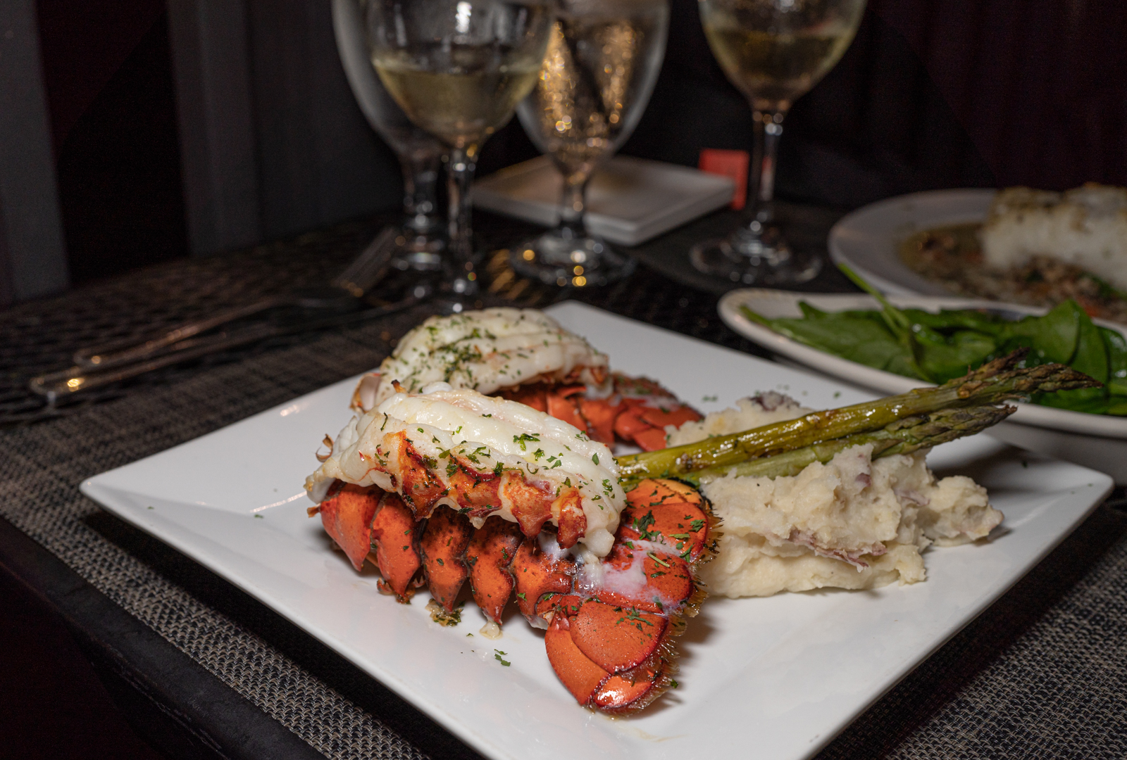 A photo at night at Oscar Penn's featuring the twin lobster tails. Asparagus rests on mashed potatoes. Behind that dish is sauteed spinach. 