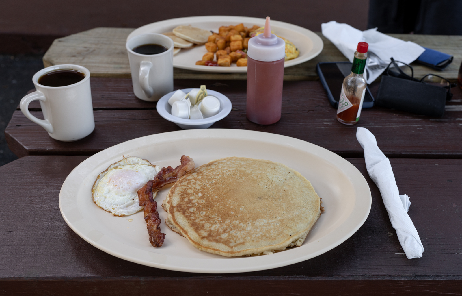 Two plates rest on a wooden picnic table. The plate in the foreground has one pancake, one piece of bacon, and one fried egg on it. There are two coffee cups behind the plate. Pats of butter on a dish, hot sauce, and ketch rest on the table with a pair of glasses and a cell phone. Hashbrowns and eggs, and small pancakes are on the other plate. 