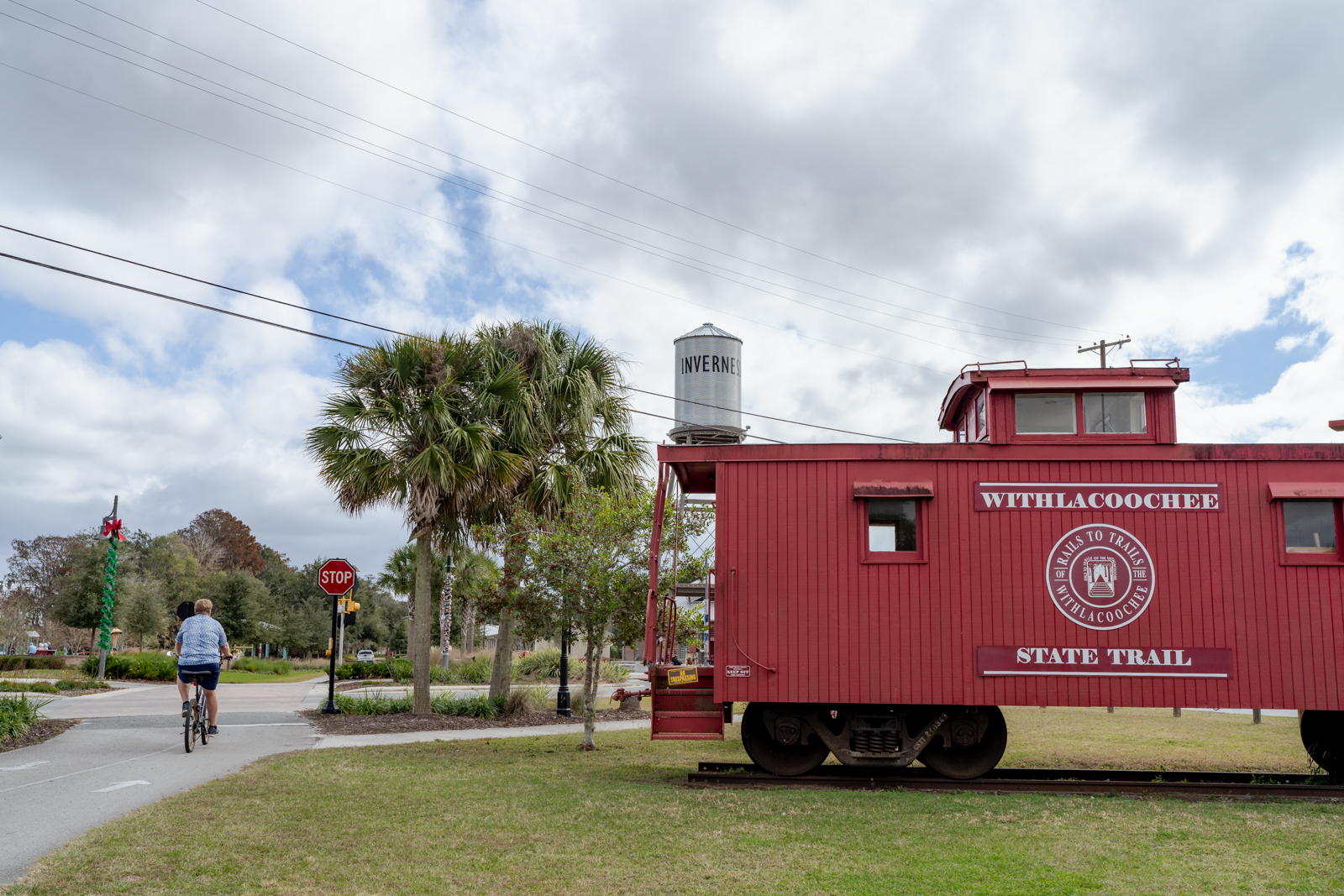 The Withlacoochee State Trail caboose is parked on the right hand side of the photo. A cyclist on the trail comes to a stop. In the background is a water tank with Inverness written on it.