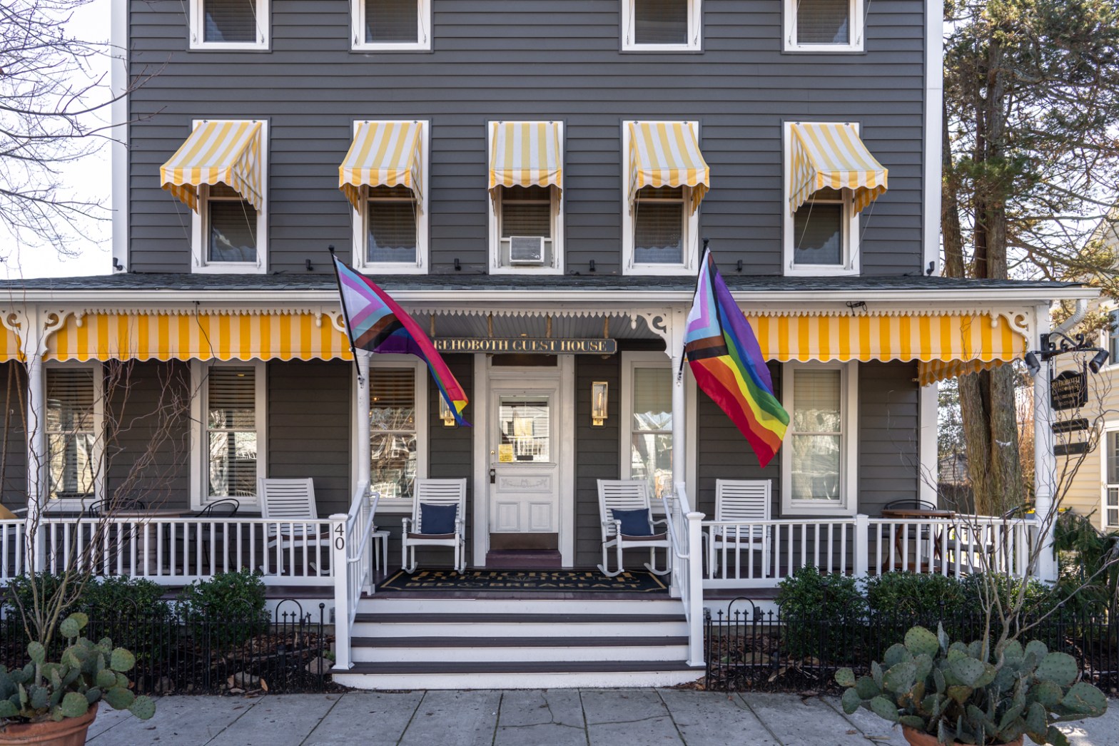 a photo of the outside of Rehoboth Guest House. The building is three stories with white windows and yellow and white striped awnings above them. There are two gay flags waving at the entrance.