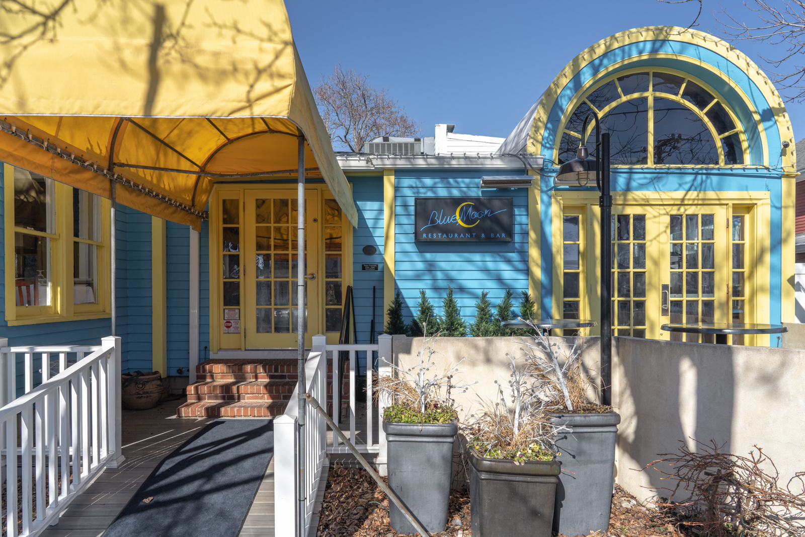 a photo of the exterior and entrance to Blue Moon Restaurant and Bar. The building is light blue with yellow doors, trim, and a yellow awning. There are three planters in front of the building. 