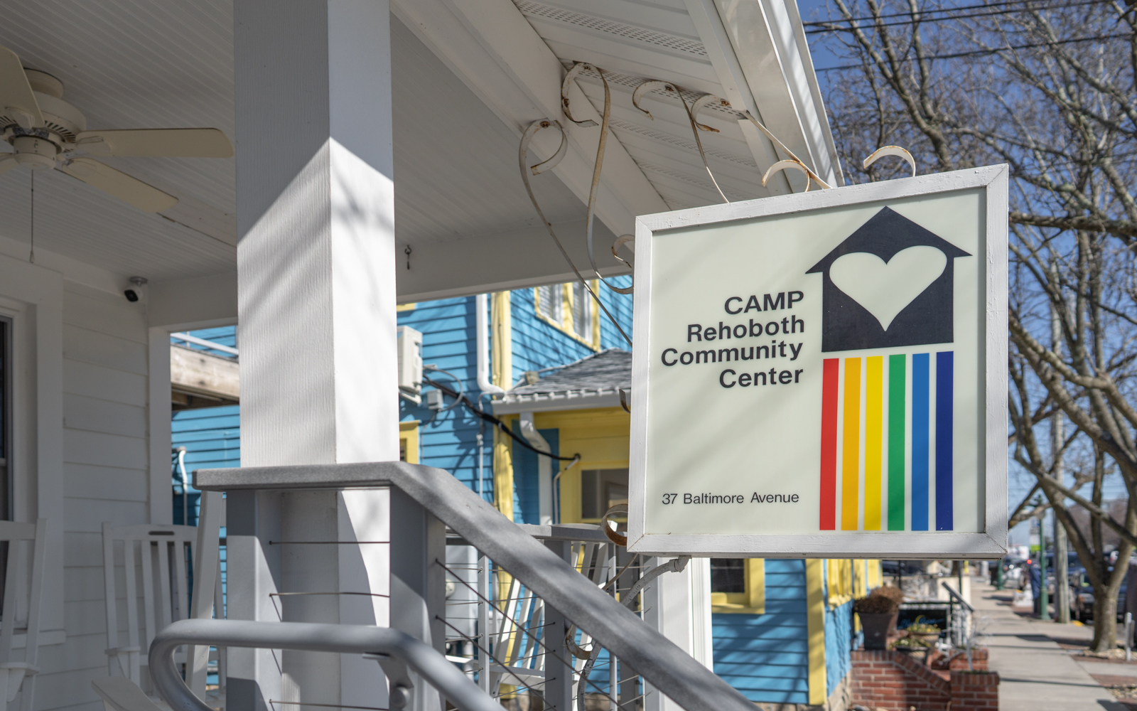 a photo of the exterior of CAMP Rehoboth Community Center. The sign is hanging from the outside of the white building. It's logo bears the LGBTQ rainbow colors. 
