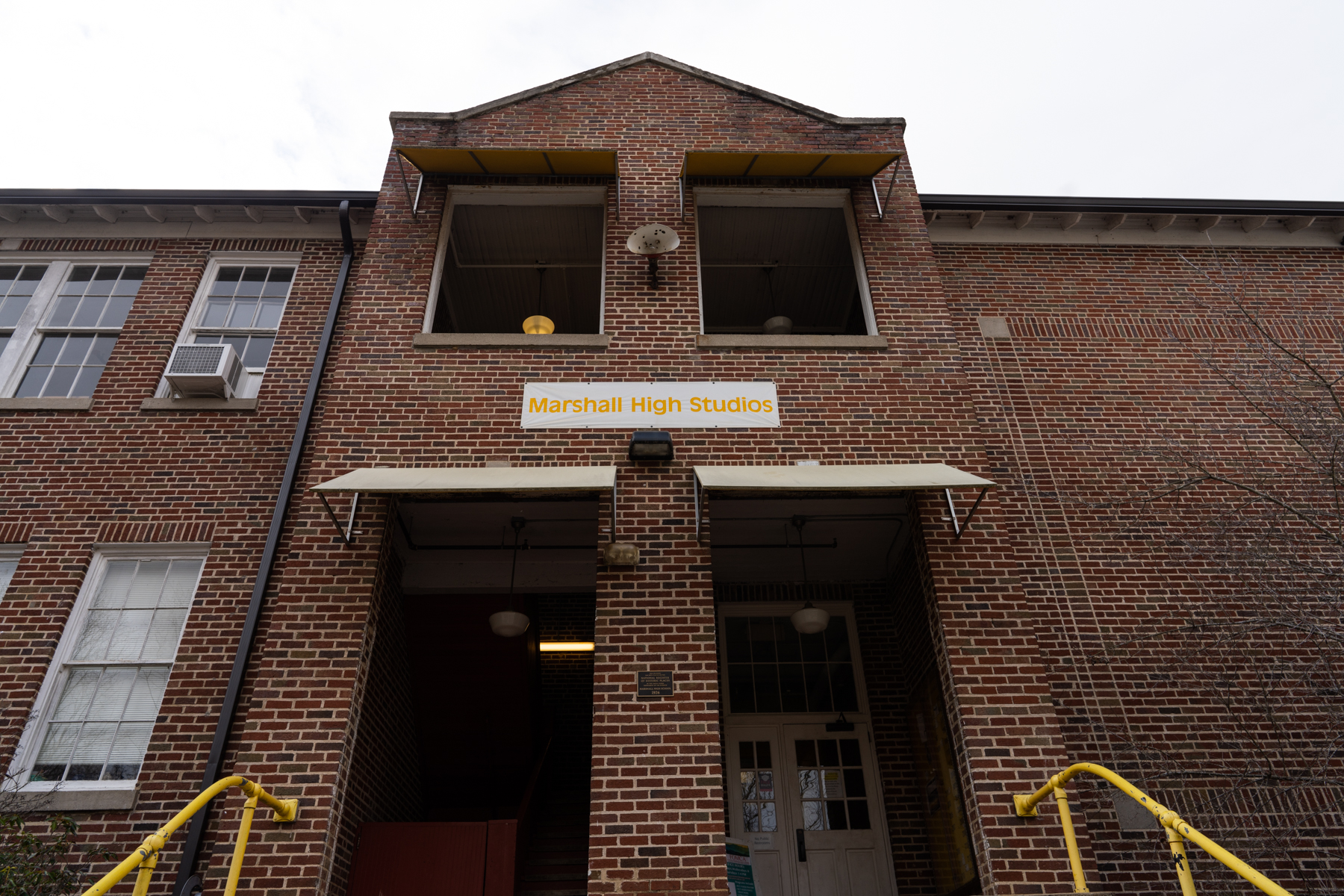 a photo of the exterior of Marshall High Studios. The building is two stories tall and made of brick. A yellow handrail goes up to the building and a white sign with yellow letters says Marshall High Studios.