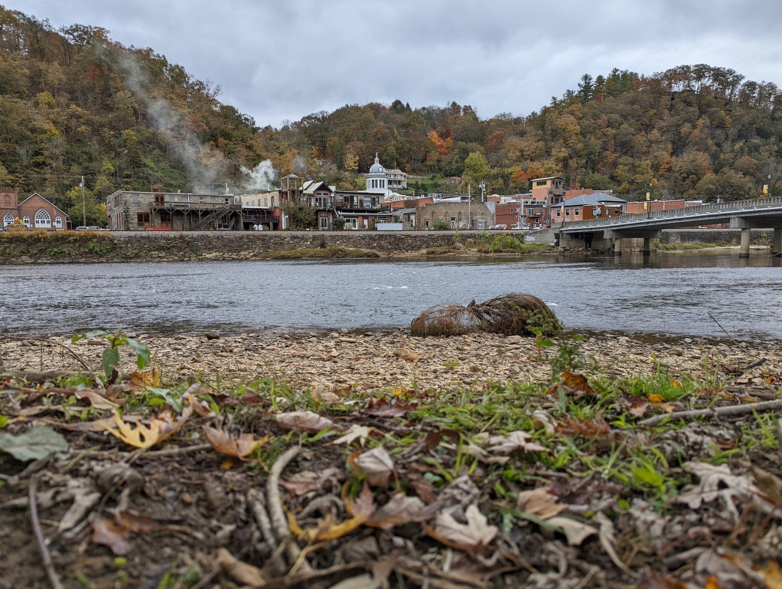 a photo of the French Broad River is the center focus of the photo. The town of Marshall, NC is in the background. Leaves are on the ground and slightly blurred out in the foreground.