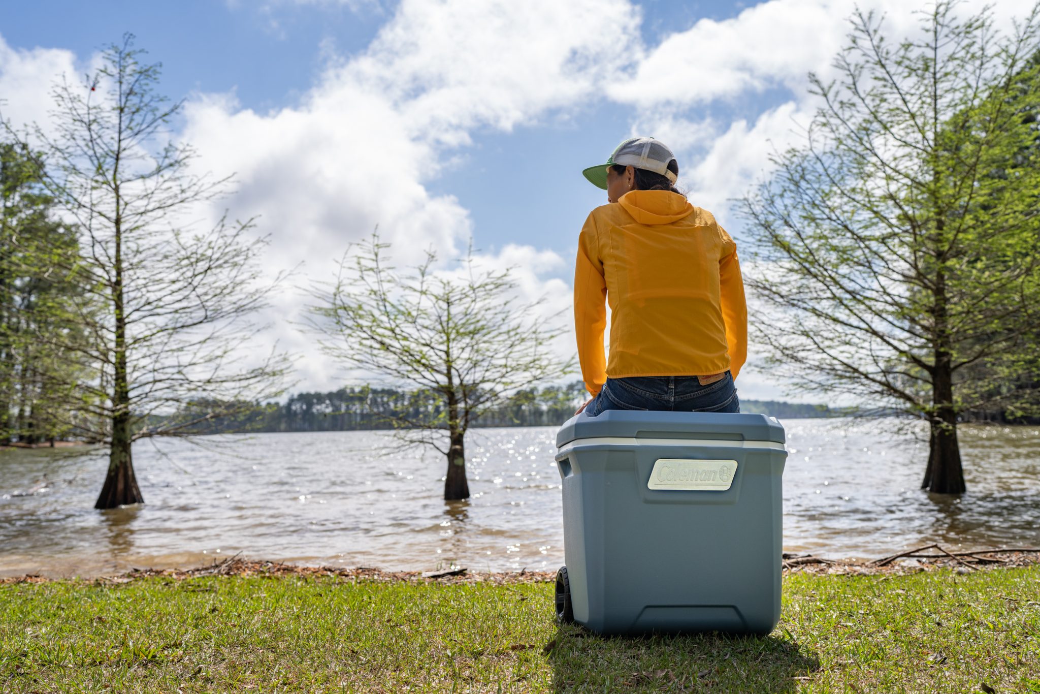 Erin McGrady is sitting on a blue Coleman Atlas 65-Quart Cooler. She is facing a lake and there are three cypress trees in the lake. She is wearing a yellow jacket and green trucker hat and jeans.