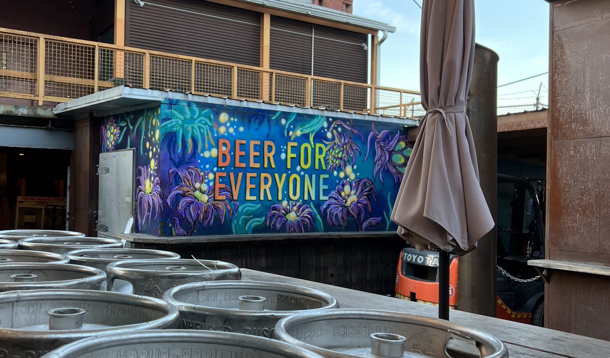 a blue mural with purple flowers and some yellow pollen that seems to be glowing has, in all caps, the words "Beer for Everyone". The words are in dark orange and as they move left to right, become yellow and then blue. There are kegs in the foreground as well as a closed tan umbrella. In the background is a building with a wooden balcony. 