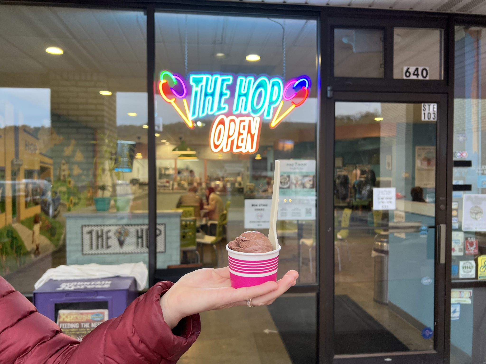 a white persons hand is holding a pink cup of brown ice cream with a spoon stuck in it. the forearm and part of the upper arm are visible in a maroon puffy coat. In the background are window panes and a glass door. There is a neon sign that's on that says THE HOP OPEN with two ice cream cones on either side.