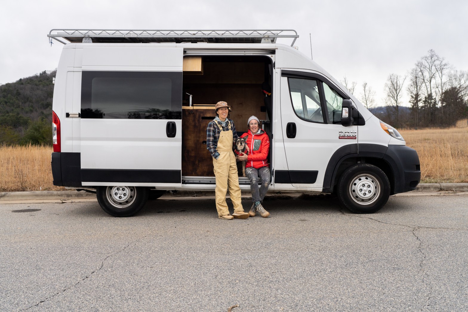Erin McGrady stands in the doorway of an open white, tall, Ram Promaster camper van. Next to her is a cute brown dog with white fur around it's mouth and eyebrows. On the other side of the dog, with her hand around the dog is Caroline Whatley. In the background is a wintry looking scene with a mountain and golden grass.