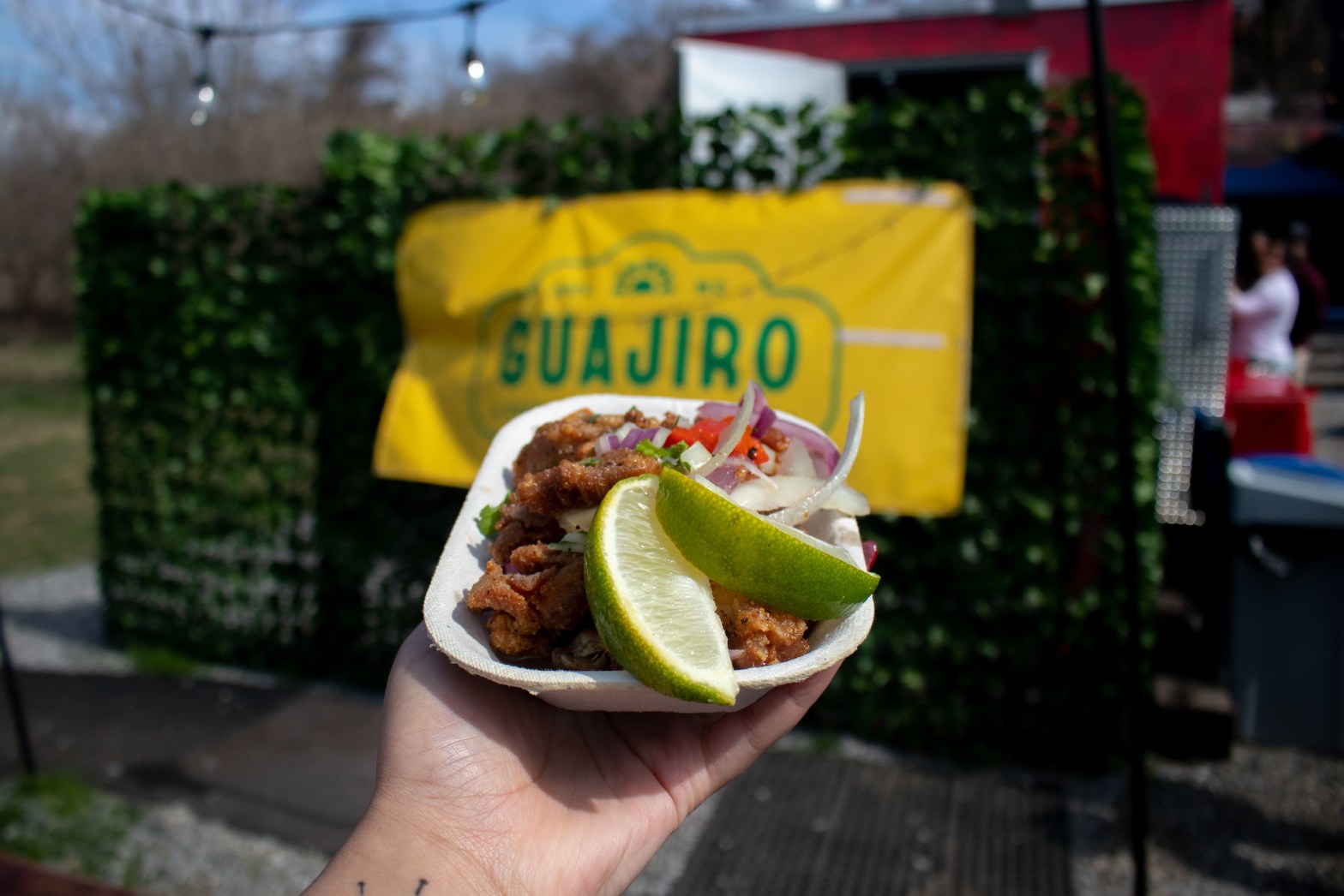 a hand is holding a white rectangular dish with some raw onion, pork, and two lime wedges. In the abckground is a fake green wall with a yellow banner on it that says Guajiro on it.