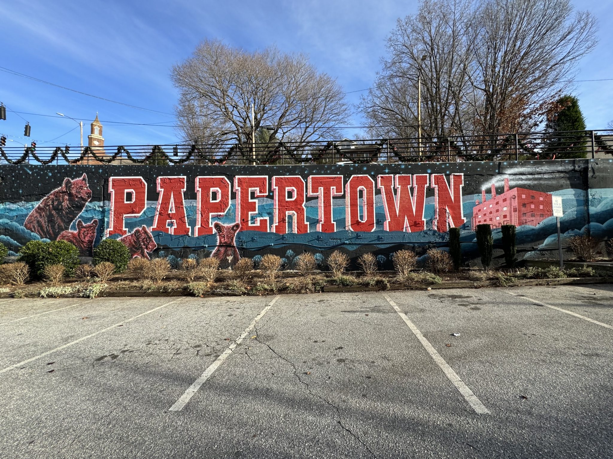 A mural painted on a wall in a parking lot reads "Papertown". The letters are in big varsity, all-caps red letters with a white outline. There are two black bears to the left of the letter "P." Some small, short landscaping is in the front of the mural. The foreground is mostly parking lot with white stripes. The background is trees without leaves and blue sky.