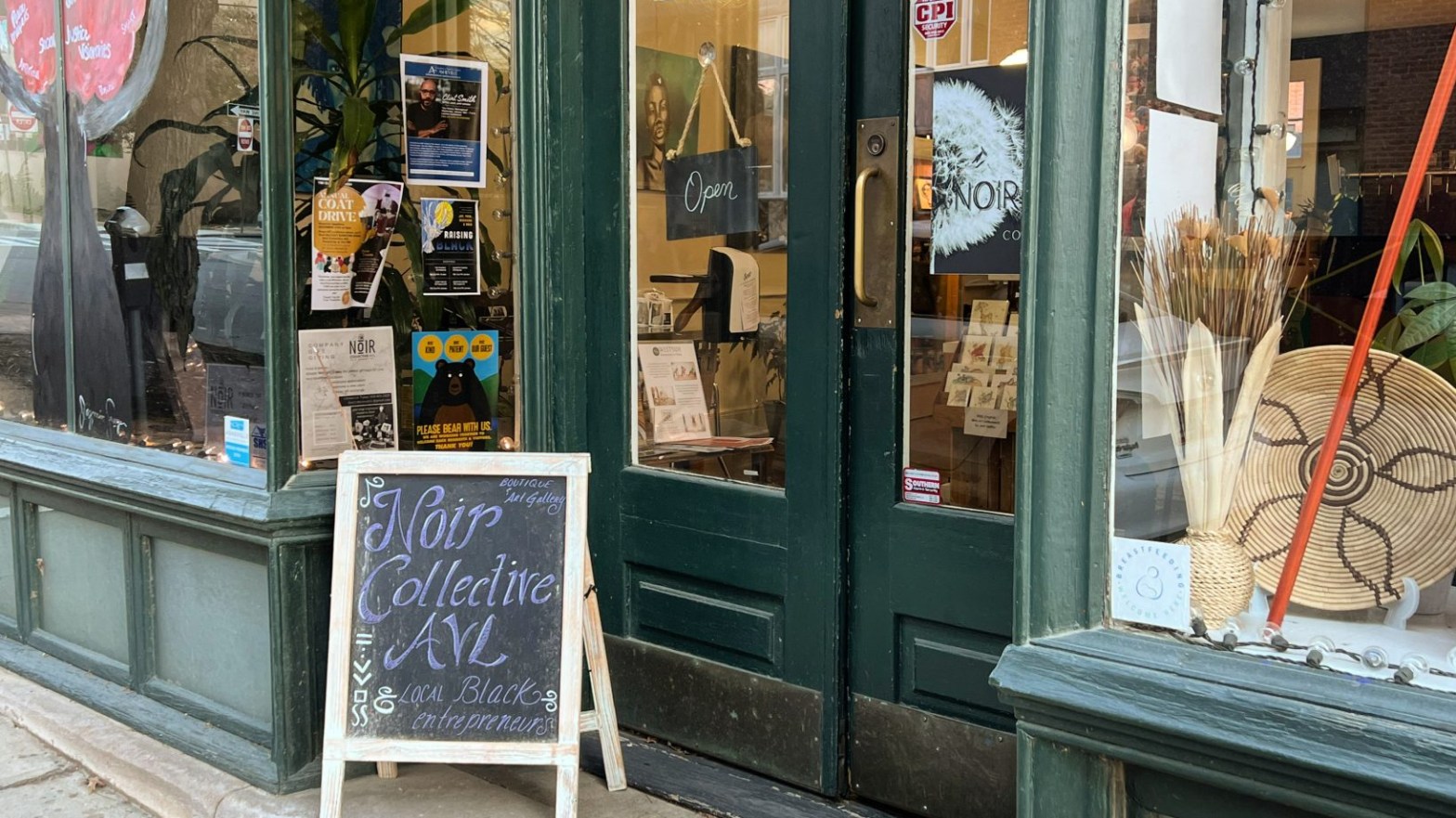 The exterior of Noir Collective AVL. The words are printed in the center of a window in white. The shop itself is green. White lights dangle from the window and it is filled with prints, books, and art.